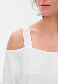 Biały top z odkrytymi ramionami, wykonany z miękkiego, zwiewnego materiału. Ramiączko ma skomplikowany, jasnobeżowy detal w formie krzyżykowego haftu, który dodaje mu charakteru.