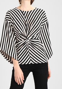 Mustat ja valkoiset raidalliset paita, jossa on solmittu yksityiskohta vyötäröllä, leveät hihat ja pyöreä kaula-aukko. Sileä kangas.