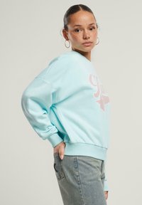Felpa azzurra chiara con scollatura rotonda e maniche lunghe. Presenta una grafica di una stella rosa e polsini a coste. Indossata con jeans azzurri chiari.