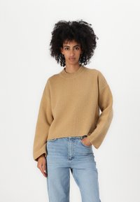 Maglione lavorato a maglia beige con collo rotondo, vestibilità comoda e maniche ampie, abbinato a jeans azzurri chiari. La texture appare morbida e accogliente.