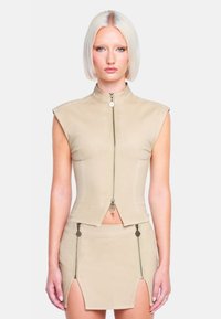 Beige verkürzte Weste mit hohem Kragen und Reißverschluss vorne, kombiniert mit einem passenden Minirock mit Seitens Reißverschlüssen und Schlitzen. Glatte Textur.