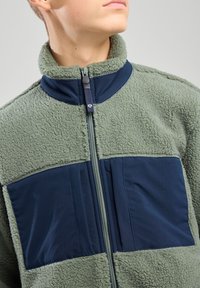 Fleece jacket featuring a light green textured fabric with dark blue nylon panels, full zipper, and high collar.