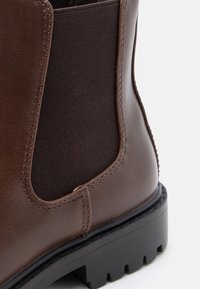 Botină din piele maro, până la gleznă, cu panouri elastice pe laturi, suprafață texturată, cusături întărite și o talpă robustă din cauciuc negru.