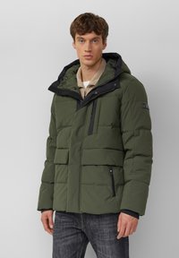 Groene geïsoleerde jas met horizontaal doorgestikt patroon, dubbele ritszakken, een zwarte capuchon en accenten. Draagt over een beige top met kraag.