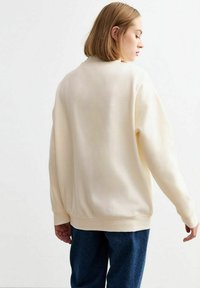 Sudadera de color crema con un ajuste relajado, puños y dobladillo de canalé. Textura de tela suave, mostrada desde la parte trasera con un ligero deslizamiento en los hombros.