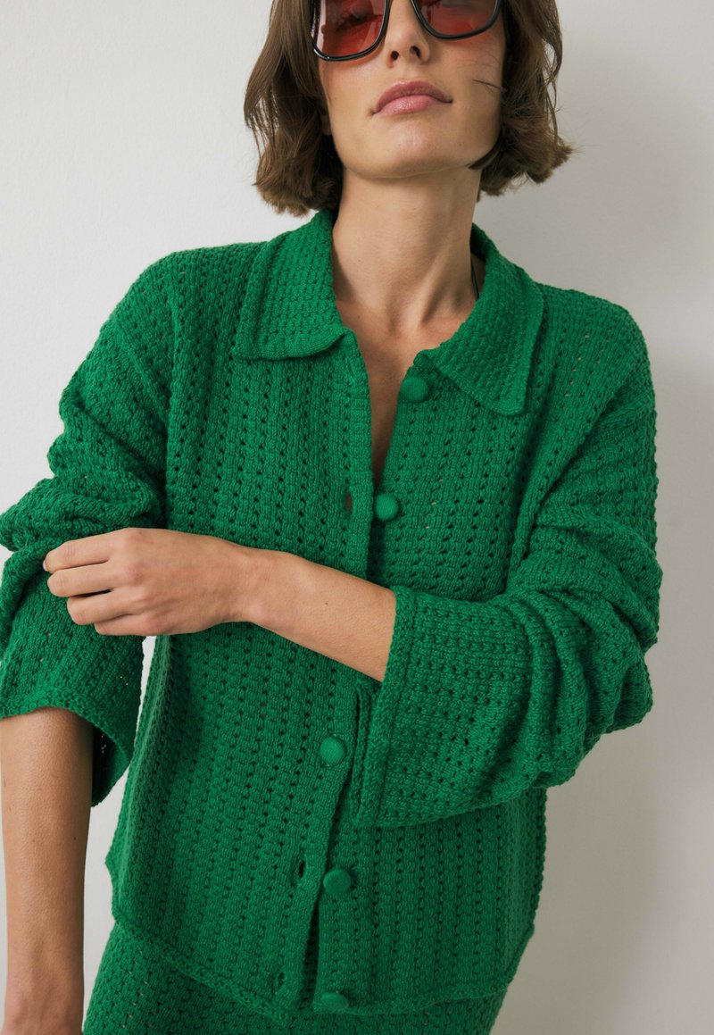 Groene gebreide cardigan met een textuurpatroon, kraag en grote knoopsluitingen. Heeft ontspannen mouwen en een comfortabele pasvorm.