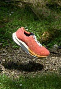Chaussure de course légère en corail avec des accents gris, tige texturée, semelle blanche et détails réfléchissants. Positionnée au-dessus d'un terrain herbeux.