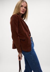 Brun fløjl blazer med en tætsiddende pasform, skrå lapeller og knapdetaljer, båret over højtaljede blå jeans.