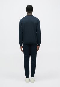 Muž v námořnicky modré mikině a kalhotách, v bílých teniskách, stojí čelem odvrácený k jednoduchému bílému pozadí.