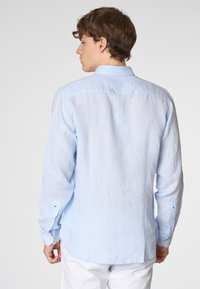Un giovane uomo, visto di spalle, indossa una camicia a maniche lunghe blu chiaro con colletto abbottonato, abbinata a pantaloni bianchi.
