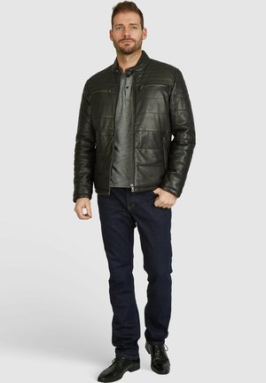 Chaqueta de cuero negro con cremallera y diseño acolchado, con dos bolsillos en el pecho, llevada sobre una camisa gris, combinada con jeans oscuros y zapatos negros.