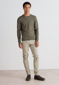 Maglione a maniche lunghe verde oliva scuro, pantaloni slim-fit beige e stivali alla caviglia marrone scuro, in piedi contro uno sfondo chiaro e neutro.