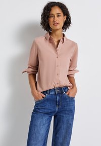 Femme aux cheveux bouclés portant une chemise rose poussiéreux à manches retroussées et un jean bleu, debout les mains dans les poches contre un mur blanc.