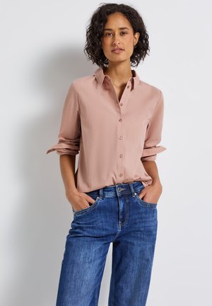 Femme aux cheveux bouclés portant une chemise rose poussiéreux à manches retroussées et un jean bleu, debout les mains dans les poches contre un mur blanc.