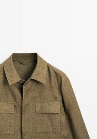 Khaki long-sleeve shirt with a collar, two chest pockets, and a textured fabric. The design features clean lines and a minimalistic style.