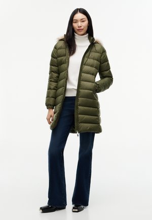 Mujer que lleva un abrigo acolchado verde oliva con capucha con borde de piel, camiseta de cuello alto blanca, vaqueros azul oscuro y zapatos negros, de pie con una mano en el bolsillo.