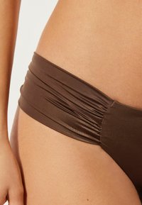 Bas de bikini marron avec une taille froncée, en matière lisse. Présente un design épuré et minimaliste, mettant en avant une silhouette ajustée.