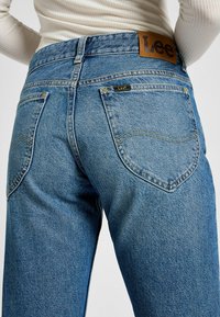 Jeans de denim azul claro con corte recto, que cuentan con bolsillos traseros, costuras amarillas y una etiqueta de cuero marrón con el logotipo de Lee en la cintura.