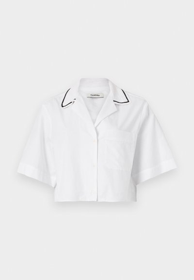 Λευκό κοντό μπλουζάκι με κοντά μανίκια, κλείσιμο με κουμπί, τσέπη στο στήθος και μαύρη ρέουλα γύρω από τον γιακά. Κατασκευασμένο από ελαφρύ, αναπνεύσιμο ύφασμα.