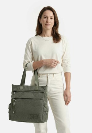 Brūniem matiem sieviete, kas tērpusies krēmkrāsas apģērbā, tur rokā lielu pelēku somu ar vairākiem kabatām ar rāvējslēdzējiem baltā fonā.