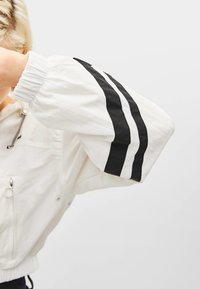 Biała krótka kurtka z elastycznymi mankietami, z dwoma czarnymi pionowymi paskami na rękawach i zapinaną na zamek kieszenią z przodu. Gładka tekstura.