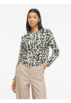 Žena s dlhými tmavými vlasmi, oblečená v bielom a čiernom kardigane s leopardím vzorom a béžových nohaviciach s vysokým pásom, stojaca s jednou rukou v vrecku.