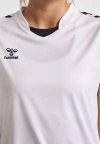 Camiseta deportiva blanca con el logo negro de hummel en el lado izquierdo del pecho, camiseta interior negra visible en el cuello en forma de V, y chevron negros en los hombros.