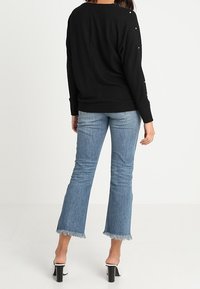 Svart tröja med knappdetaljer på ärmarna, tillsammans med blå utsvängda jeans med fransar i nederkant, och svarta klacksandaler.