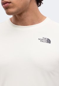 Κοντινό πλάνο ενός ατόμου που φοράει λευκό t-shirt με μικρό μαύρο λογότυπο The North Face στο πάνω αριστερό μέρος του στήθους.