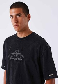 Zwarte oversized T-shirt gemaakt van versleten stof met een centraal logo en verspreide gaten. Korte mouwen met een iets wijde halslijn.