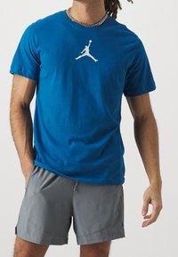 Blå bomulls T-shirt med korta ärmar som har en vit jump man-logotyp på bröstet; bärs med ljusgrå shorts och ett silverfärgat kedjehalsband.