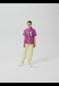 Fille debout portant un t-shirt violet avec les lettres "B C" et un pantalon jaune, des baskets blanches, sur un fond blanc uni.