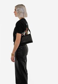 Schwarze Lederhandtasche mit strukturierter Form und verstellbarem Schulterriemen, gezeigt an einer Person, die ein grafisches schwarzes T-Shirt und Lederhosen trägt.