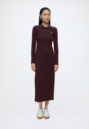 Femme aux cheveux longs, raides et noirs, portant une robe polo bordeaux cintrée à manches longues et des baskets blanches, debout devant un fond blanc uni.