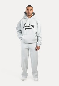 Felpa grigia chiara con logo nero "Smilodox", tasca frontale, abbinata a pantaloni della tuta coordinati. Tessuto morbido, vestibilità comoda, design minimalista.