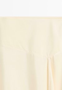 Crèmekleurige stoffen sectie met diagonale naad en lichte plooien, met textuur en zachte vallende details.