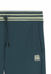 Teal joggers with a ribbed light green waistband, adjustable drawstring, and an embroidered logo of a bull with "HUGO" below.