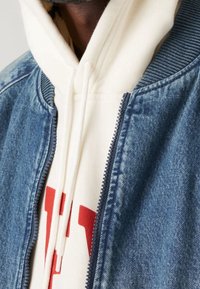 Denim bomberjacka med ribbad krage, blå tygtextur, uppknäppt för att avslöja en krämfärgad hoodie med en djärv röd tryck.