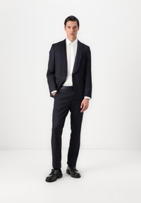 Tmavo modrý oblek so slim fit strihom, sakom na jedno gombík a zodpovedajúcimi nohavicami, kombinovaný s bielou košeľou a čiernymi topánkami. Hladká textúra látky.