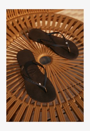 Sorte flip-flops med teksturerte såler og tynne stropper, prydet med en liten dekorativ edelsten ved sammenføyningen, plassert på en rund, flettet overflate.