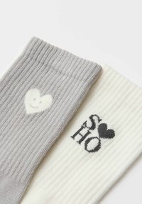 Calzino grigio a coste con motivo a cuore bianco, abbinato a un calzino crema a coste con la scritta "SOHO" e un dettaglio a cuore nero.