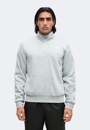 Mann mit dunklen Haaren trägt einen hellgrauen Pullover mit Viertel-Reißverschluss und schwarze Hose, steht frontal vor einem einfarbigen hellen Hintergrund.