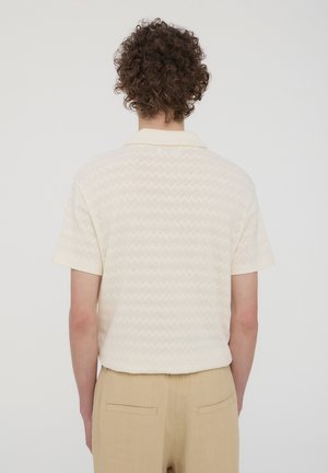 Polo a maniche corte di colore crema con un motivo a zigzag testurizzato, dotato di un colletto classico e una vestibilità rilassata, abbinato a pantaloni beige.