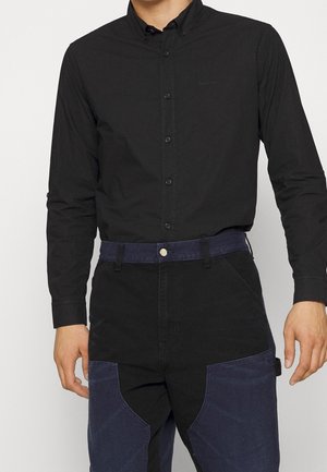 Camicia nera con bottoni, dal taglio slim e maniche lunghe, abbinata a pantaloni scuri multicolore con grandi tasche frontali e una cintura blu.