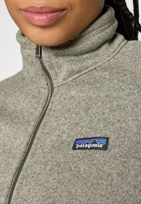 Gray zip-up fleece jacket with a textured knit fabric and a Patagonia logo patch featuring blue, red, and black accents on the left chest.