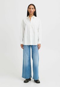 Camicia bianca a maniche lunghe con vestibilità rilassata, abbinata a jeans a gamba larga di un blu chiaro e stivali platform neri. Tessuto morbido e design classico.