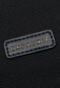 Tissu noir texturé avec un patch en cuir noir cousu affichant le mot "EXPATRIÉ" en lettres dorées.
