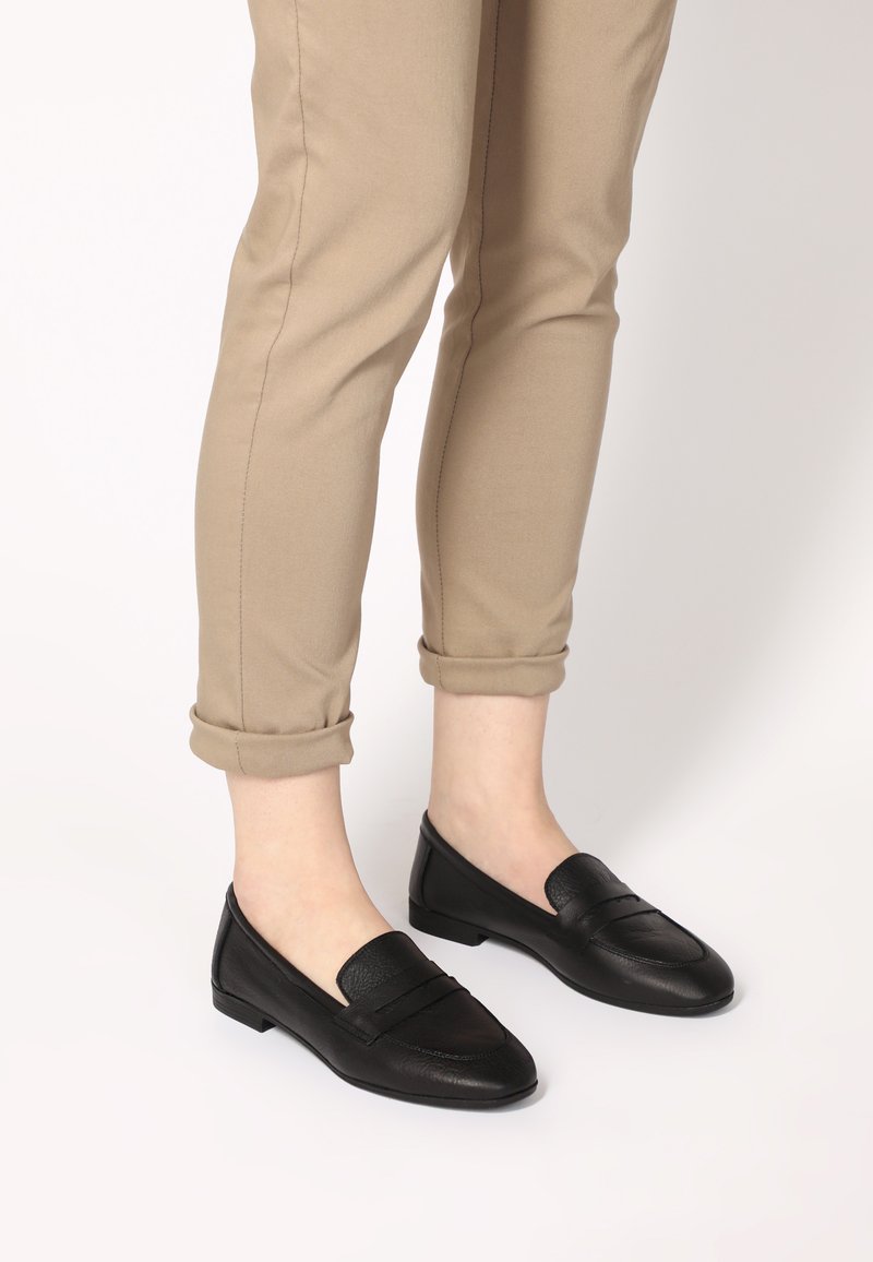 Sorte læder loafers med flad sål, der har en glat tekstur og en frontrem detalje, båret med opholdte beige bukser.