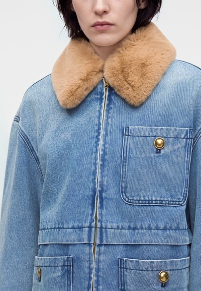 Femme portant une veste en jean bleu clair avec un grand col en fourrure beige et des poches à boutons dorés, vue des épaules au milieu du torse.