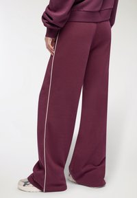 Osoba ubrana w szerokie bordowe spodnie z białymi bocznymi paskami oraz pasującą bluzę, w zestawieniu z białymi i szarymi sneakersami.